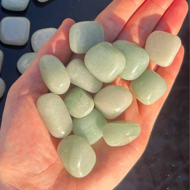 A hand overflowing with a collection of smooth, polished green aventurine tumble stone crystals, showcasing soft mossy green shades, shimmering surfaces, and glassy textures to demonstrate physical pocket sizing scale for Heart Chakra alignment.