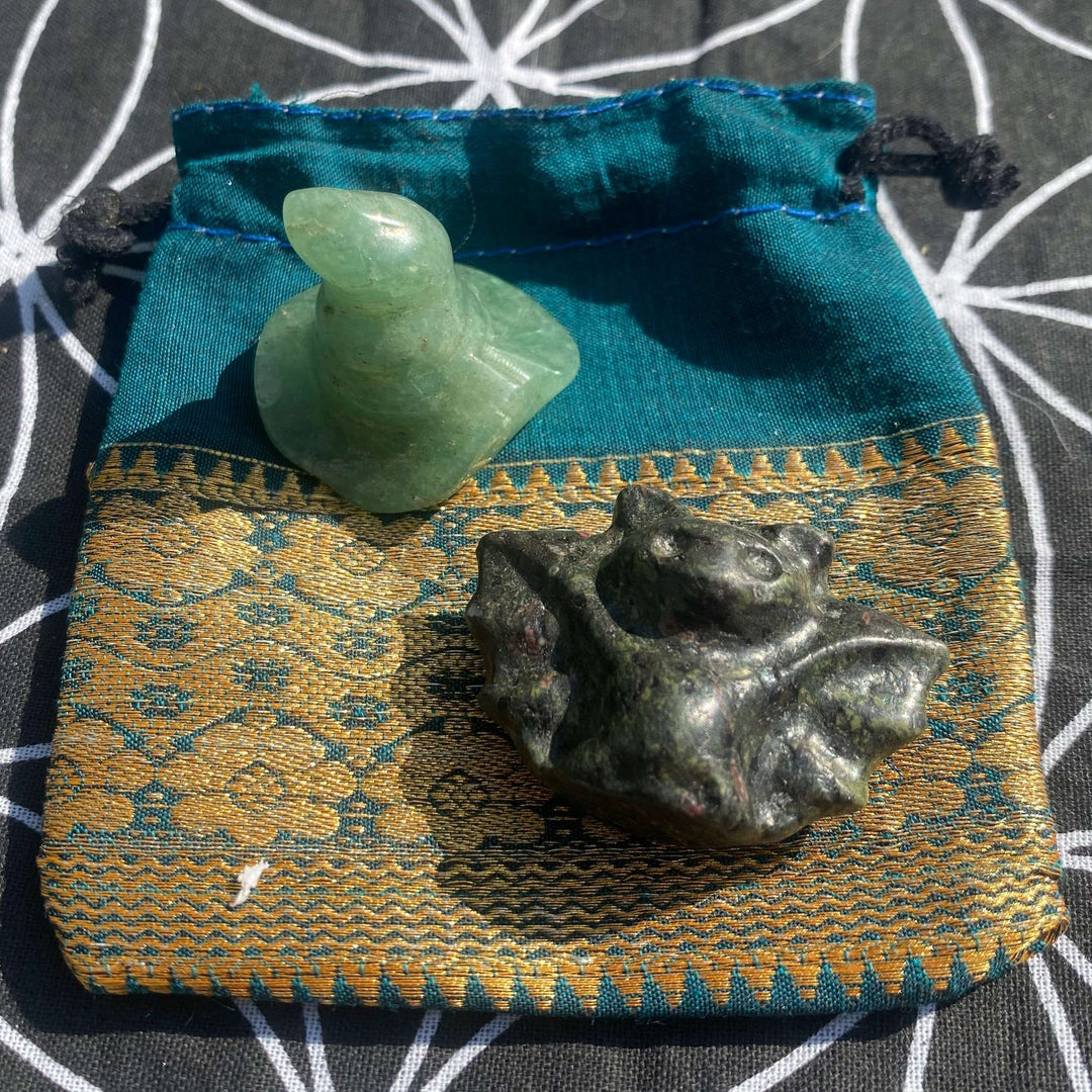 A Halloween crystal gift set featuring a hand-carved mint green Aventurine witch's hat and a dark forest-green Moss Agate bat figurine arranged on a blue and gold jacquard drawstring pouch against a geometric backdrop.