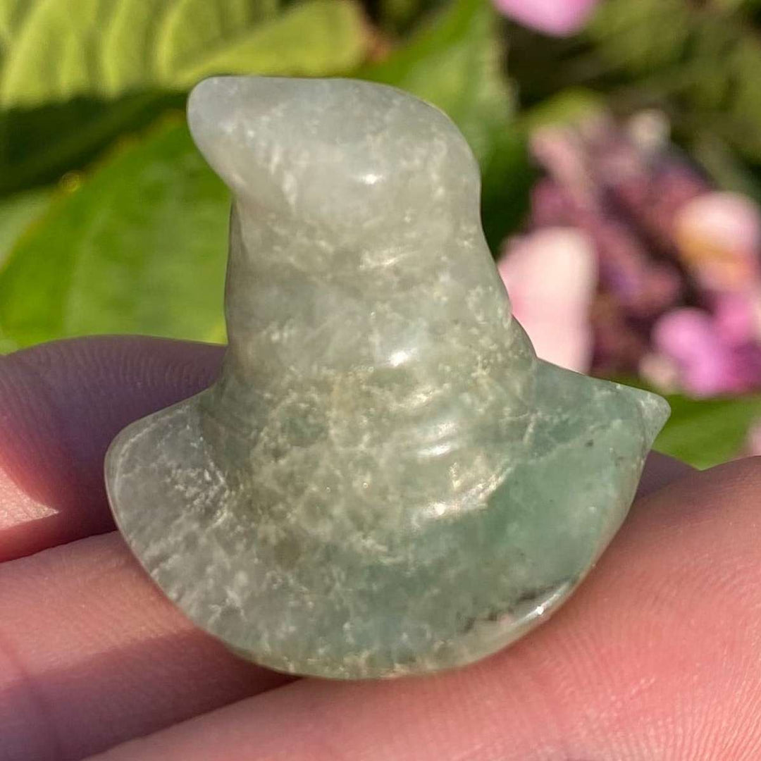 A close-up view of the small polished Green Aventurine Witch's Hat carving held in a hand. The translucent pale green crystal highlights natural mineral textures and a tiny chipped edge along the rim, captured against a blurred garden backdrop.