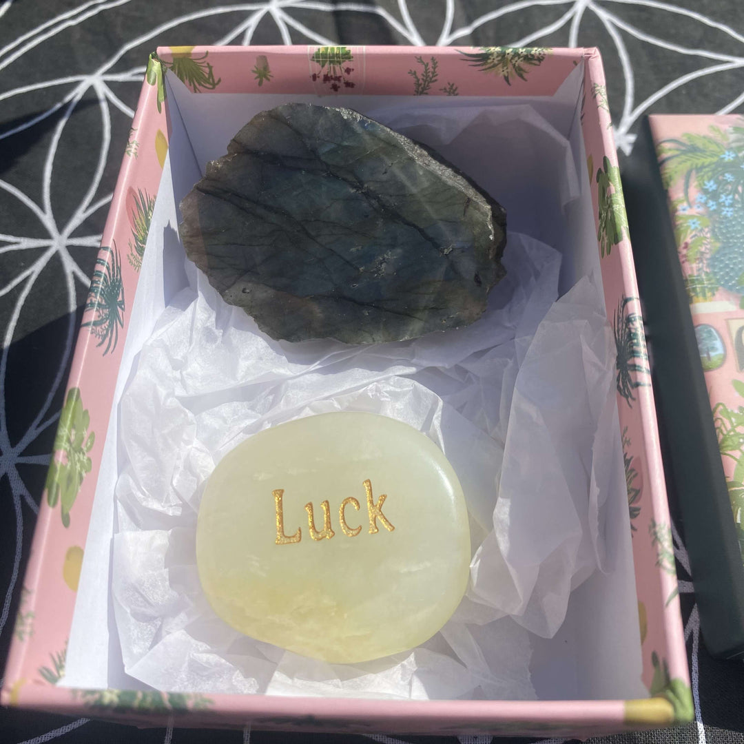 An open pink patterned crystal gift box lined with soft white tissue paper, displaying a small collection indoors. Inside sits a rough, smoky grey raw labradorite crystal chunk alongside a smooth, oval green jade "LUCK" thumbstone engraved with elegant gold lettering.