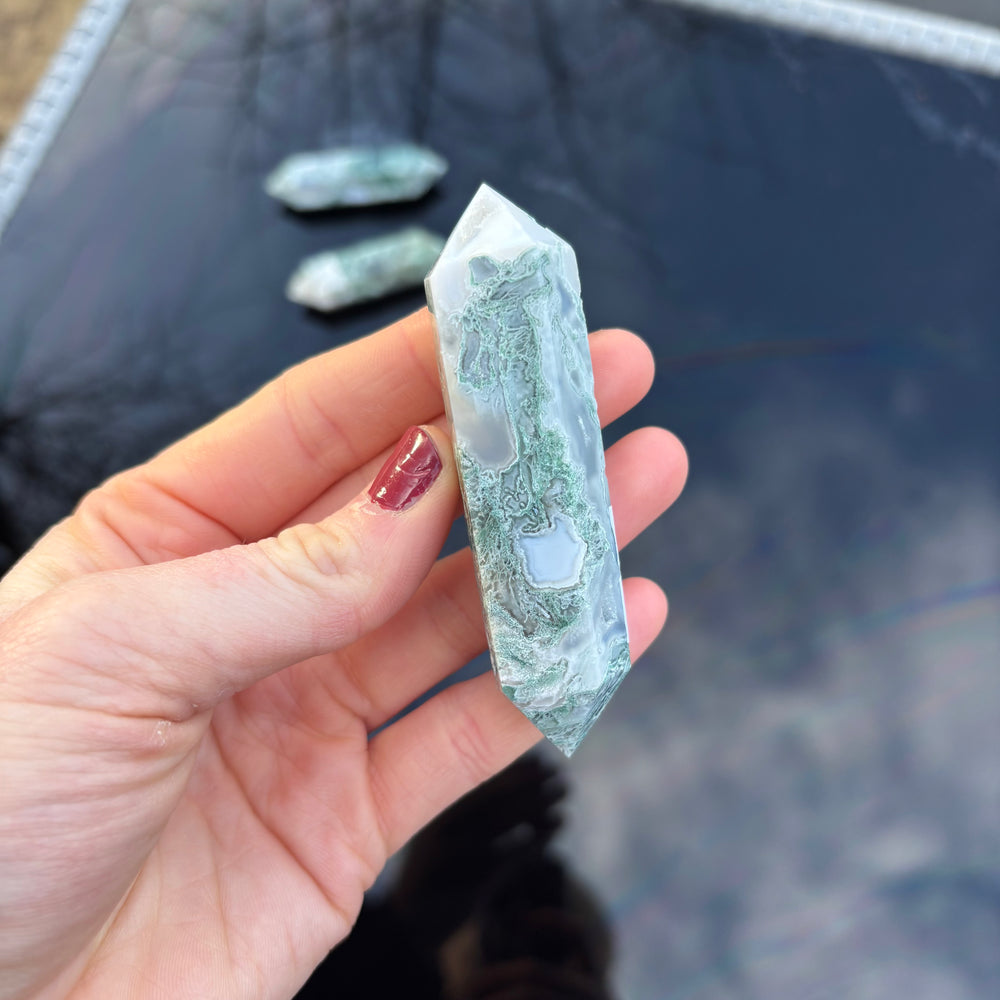 A hand holding a slender green Moss Agate dual-pointed wand crystal against a dark outdoor reflective backdrop, displaying its natural translucent patterns.