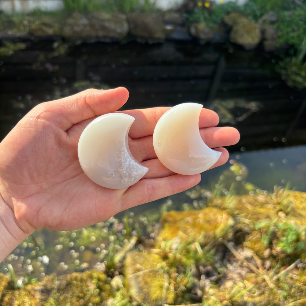 A hand holding two 6cm grey agate crescent moon crystal shapes at a distance outdoors, demonstrating the physical sizing scale relative to a palm and how the gentle celestial tones complement a mindful wellness or meditation altar setup.