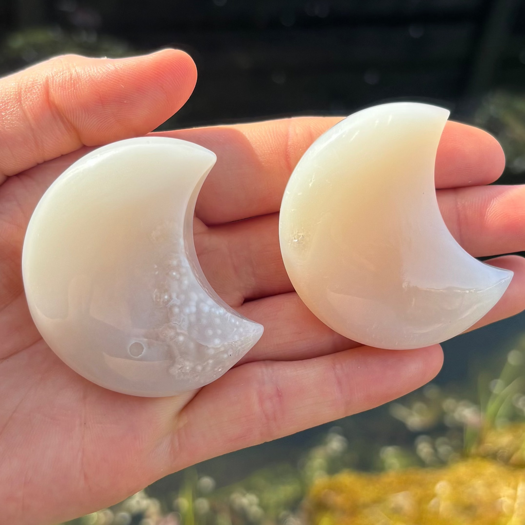 An open hand holding two polished grey agate half moon carvings outdoors, showcasing the smooth crescent silhouette, soft banded layers, and subtle internal druzy sparkle catching bright natural sunlight over a blurred garden backdrop for Root Chakra grounding.