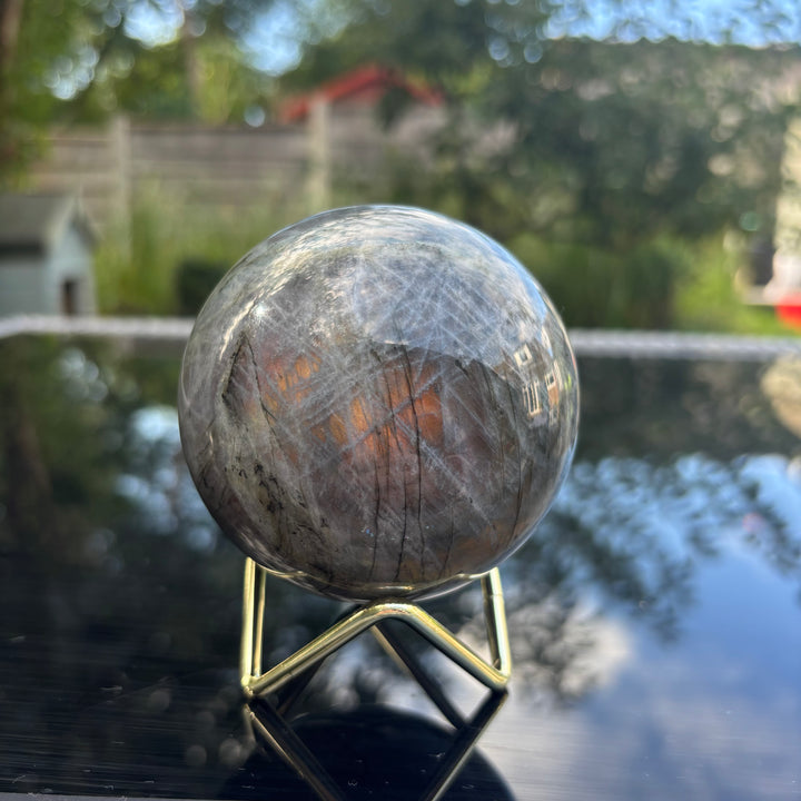 An outdoor shot of the grey labradorite 696g crystal sphere sitting on its metallic stand against a lush garden backdrop, catching natural daylight to highlight its role as a powerful energetic protective shield.
