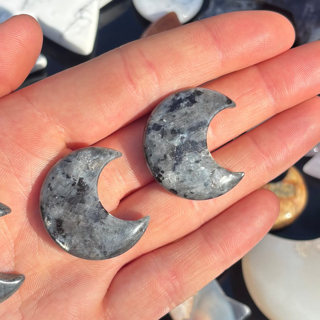 A close-up view of two carved Larvikite half moon pieces angled softly across a palm in an outdoor setting. The substantial, texturised mineral zones and subtle starry shimmers provide a protective blanket of safety, making them a tender gift to clear the mind and deflect heavy energy.