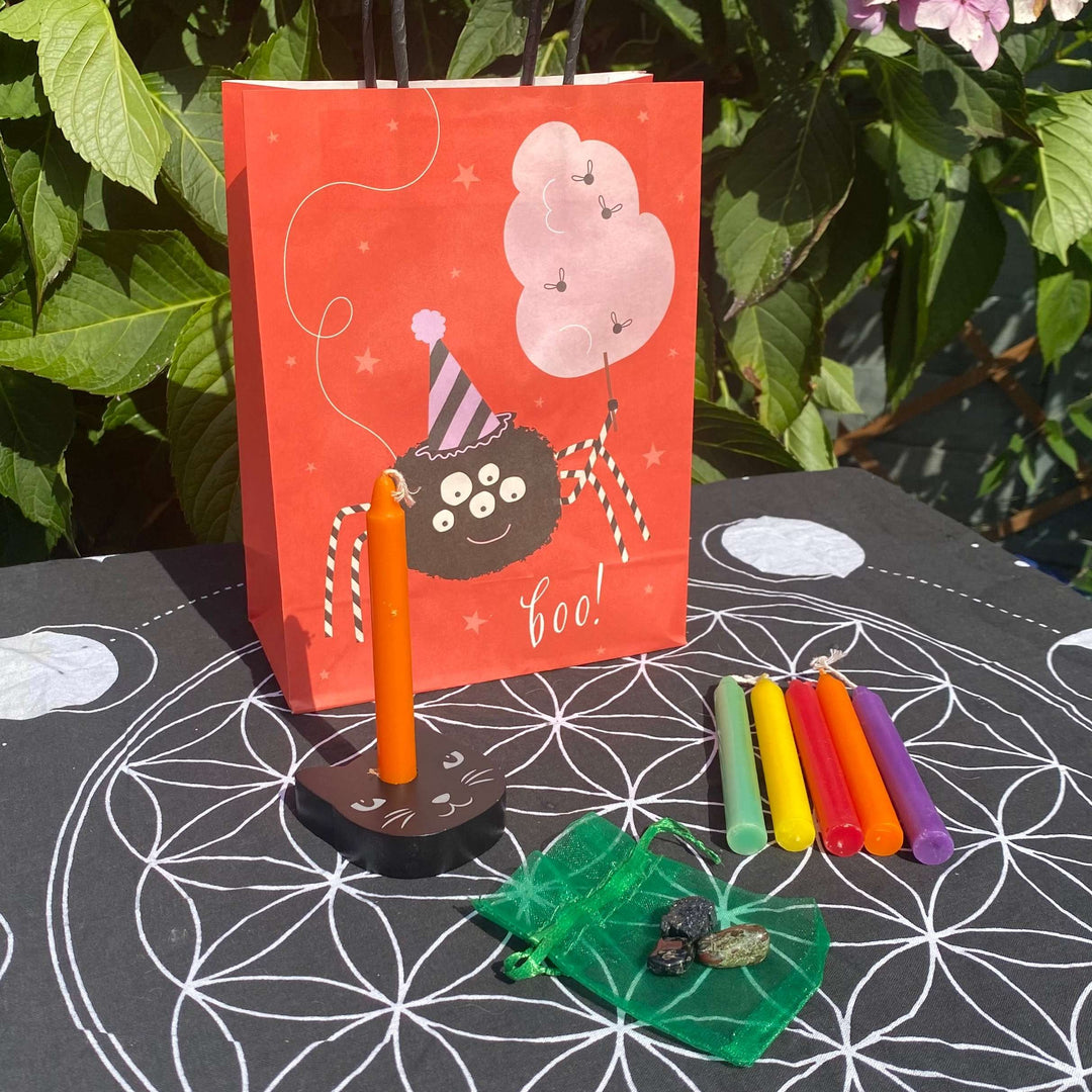 A Halloween crystal and ritual candle gift set on an altar mat, featuring an orange spider gift bag, a black cat candle holder with an orange chime candle, five ritual candles, and pocket stones in an organza pouch.
