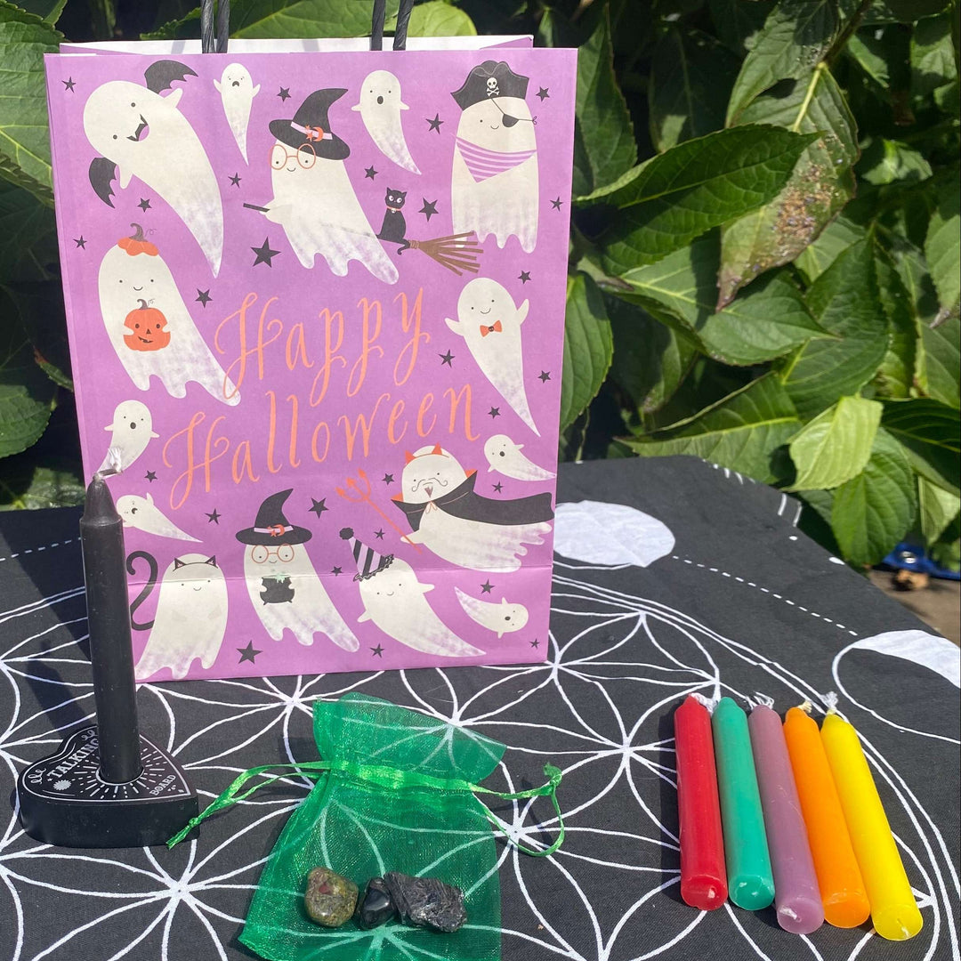 Features an overhead shot of the gift set on a Flower of Life mat, including a ghost-illustrated pink gift bag, black candle holder, five spell candles, and pocket stones in an organza pouch