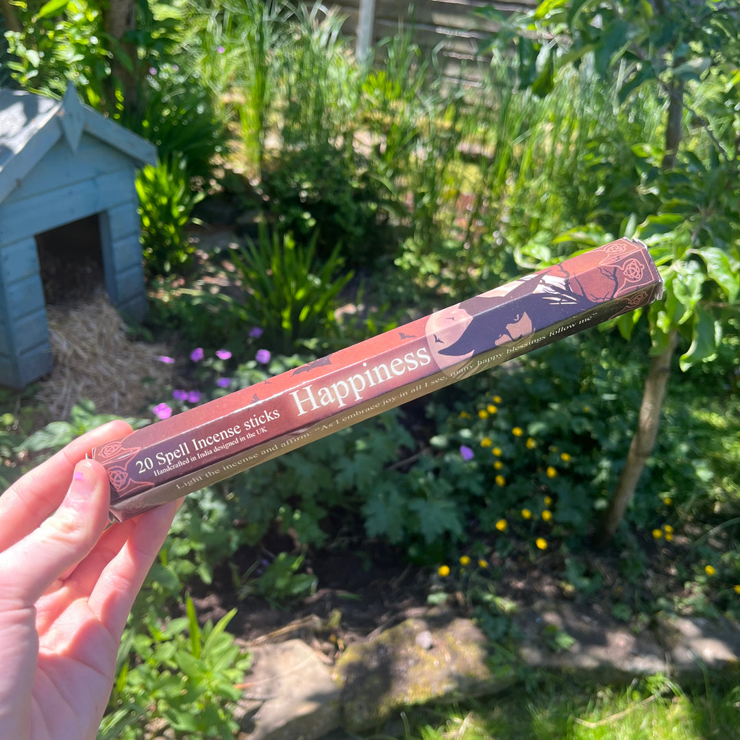 A hand holding the thin pack of happiness spell incense sticks at a distance outdoors, highlighting the sweet lavender, orange blossom, and thyme fragrance companion styled against sunlit garden greenery to welcome a bright wave of joy.