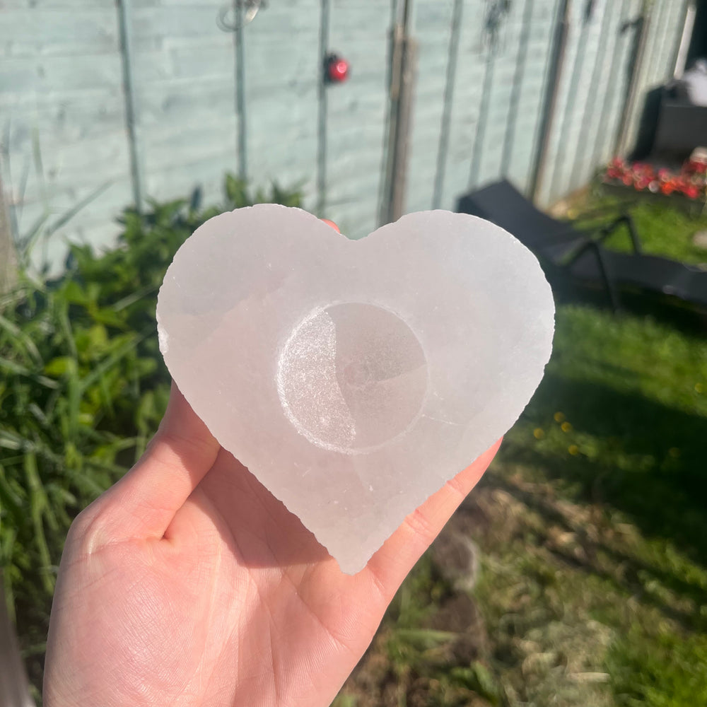A hand holding the thick heart-shaped selenite candle holder at a distance outdoors, demonstrating its approximate 10cm by 12cm physical sizing scale and soft pearlescent glow against a blurred garden lawn setting.