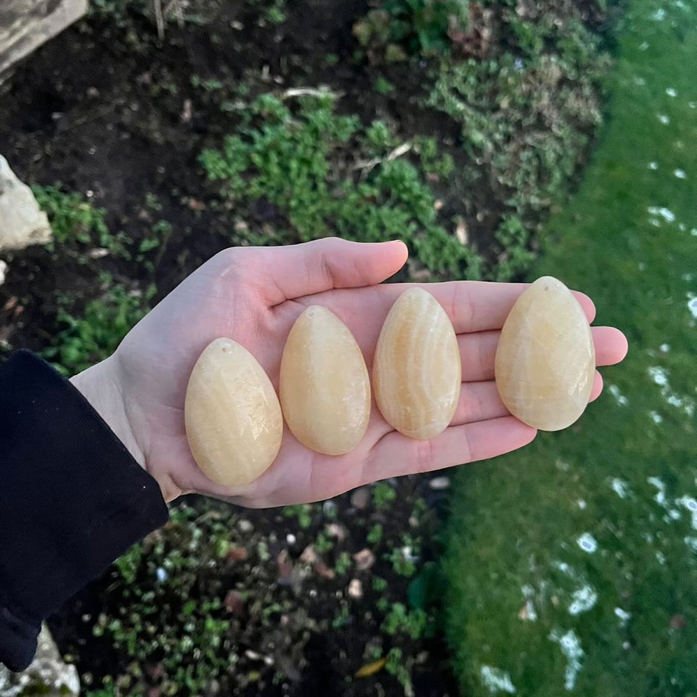 A hand holding four honey-coloured yellow calcite top-drilled teardrop crystals in a row against a green garden background