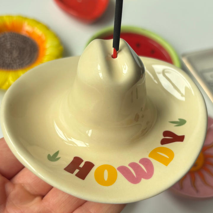 A direct close-up shot of a glossy cream ceramic incense stick holder shaped like a cowboy hat, held gently in a hand. The wide, upward-curved brim acts as an ash catcher and features multi-coloured block lettering spelling "HOWDY" flanked by tiny green cacti, with a black incense stick burning from the top hole of the hat&