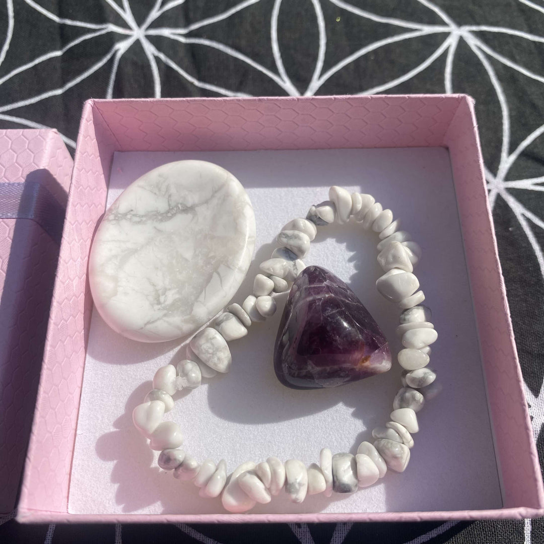 A top-down view looking into an open, square light pink box displaying the premium mindfulness crystal gift set. Inside, a textured white Howlite chip bracelet encircles a deeply polished triangular purple Amethyst tumble stone, positioned next to a smooth oval Howlite thumb stone on a clean white liner.