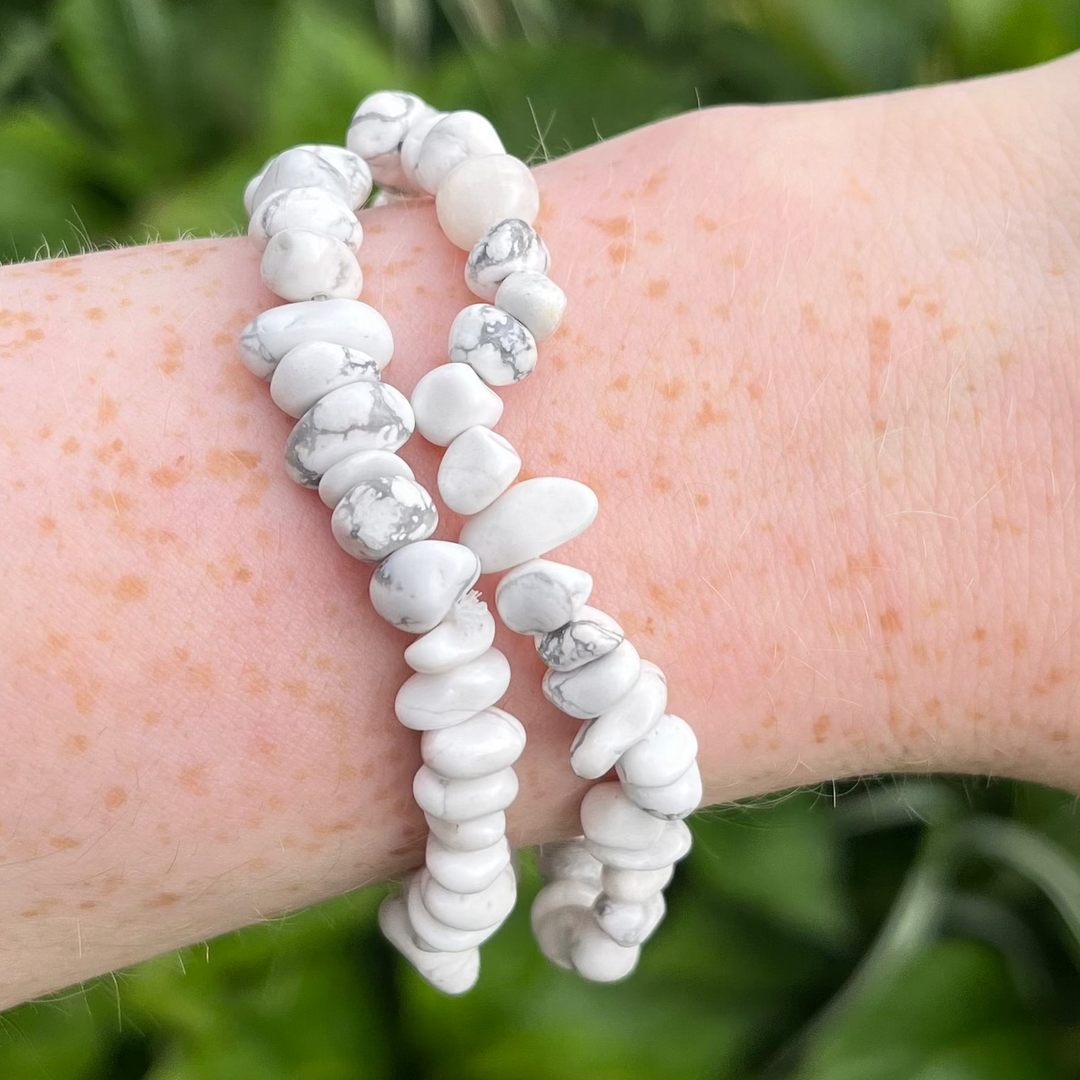 A close-up view of a wrist wearing two howlite crystal chip bracelets layered together outdoors, showcasing the polished, organic white gemstone chips with unique grey marbling patterns against a blurred green foliage background for Crown and Root Chakra balance.
