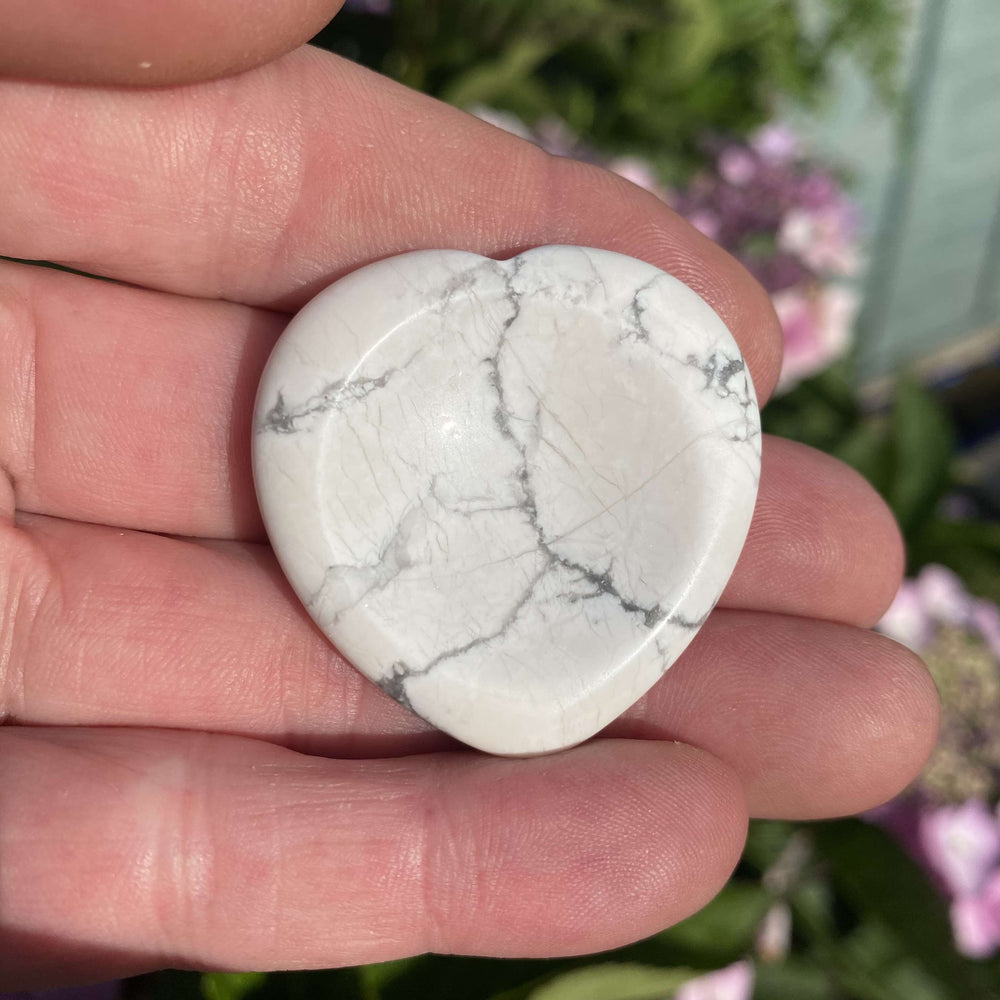 An open palm holding a small howlite crystal heart outdoors to show the size scale against a background of garden foliage.