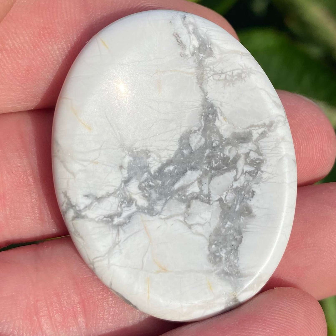 A close-up view of a polished white howlite oval worry stone held between fingers showing its velvety texture and prominent grey veining.