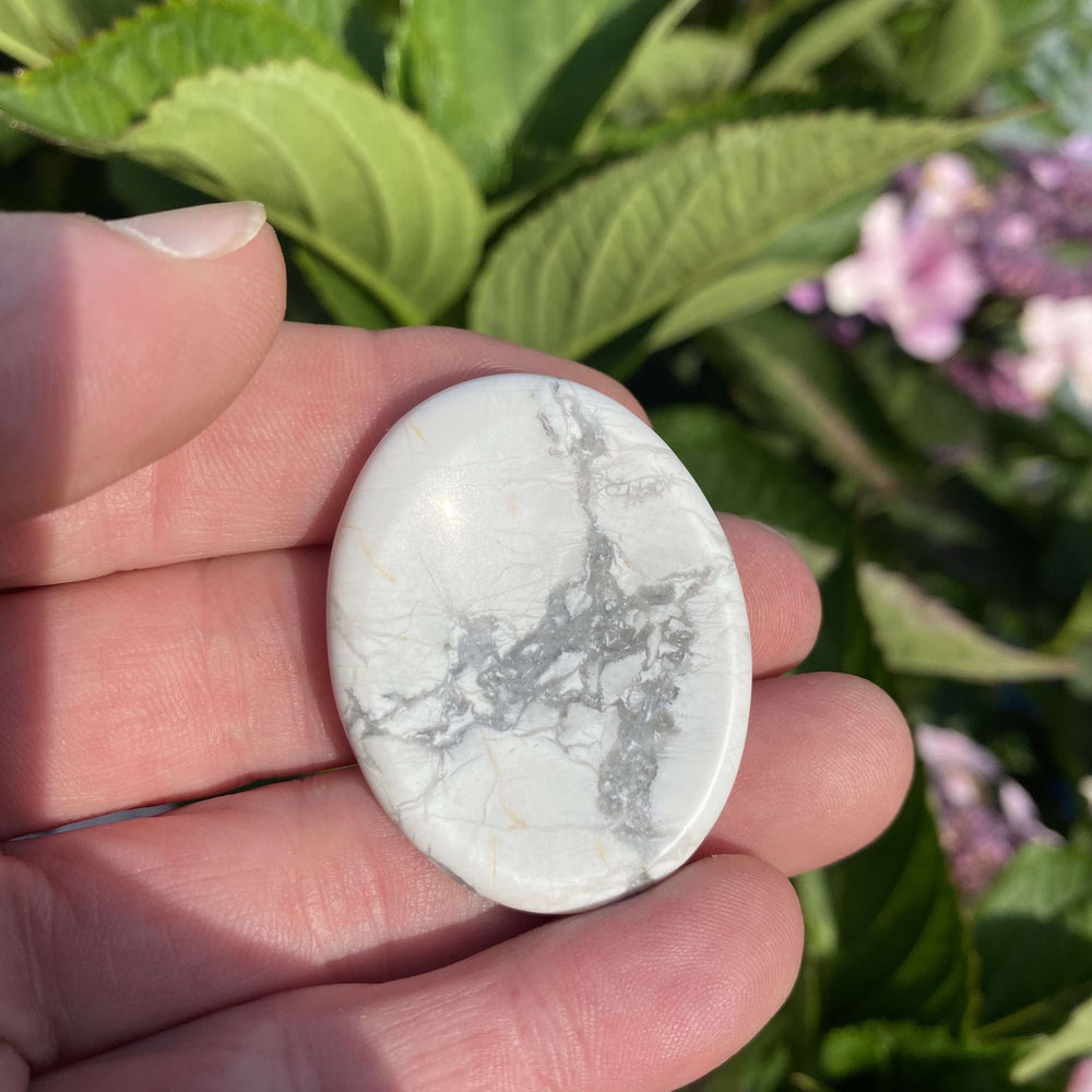 An open palm holding the smooth howlite pocket worry stone outdoors to demonstrate size scale against a background of green garden foliage.