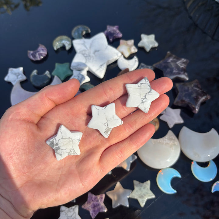 A hand presenting three ivory white howlite star carvings at a distance against a wide background of assorted pocket shapes, illustrating the physical sizing scale of these small cosmic talismans designed to bring a stable aura of deep peace.
