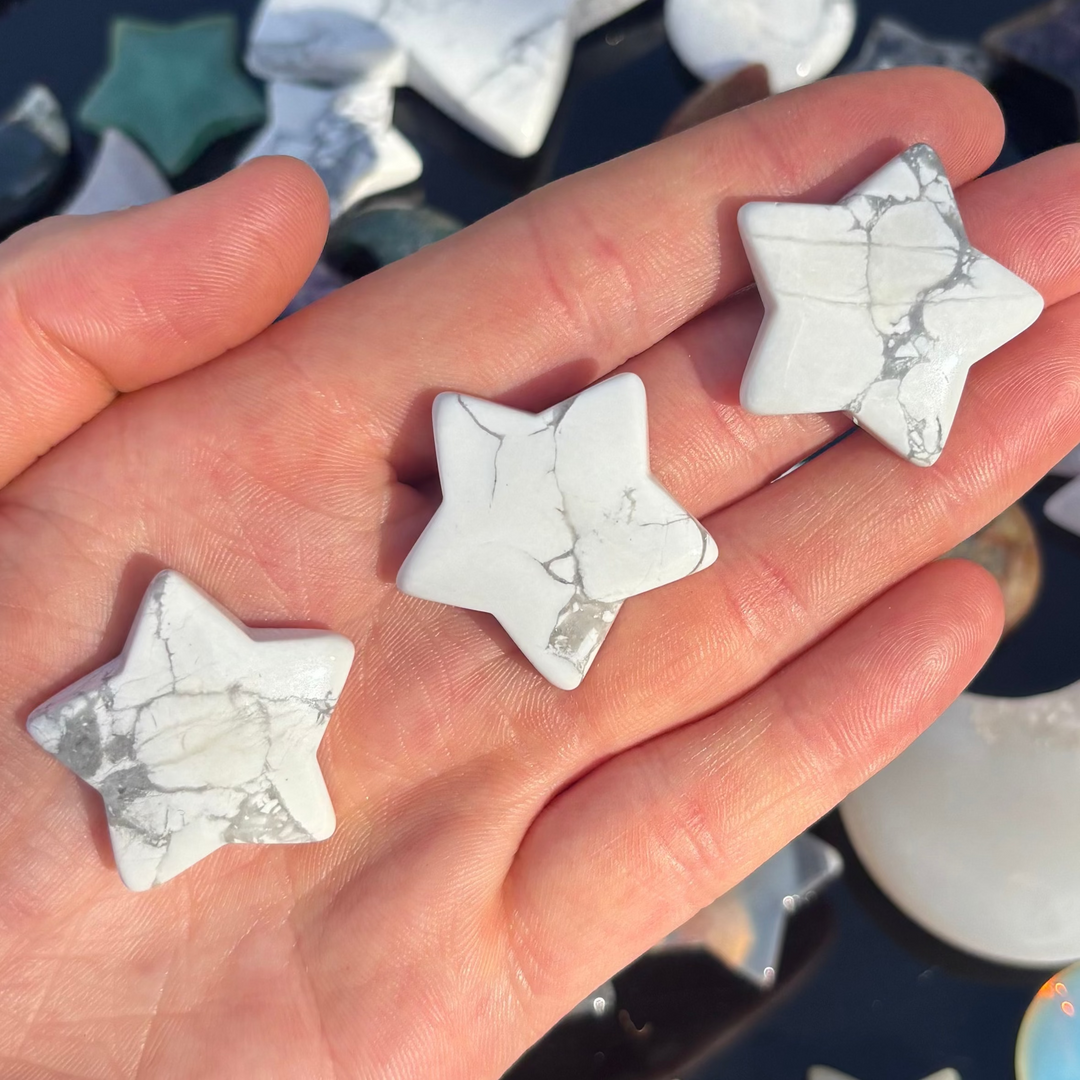 An open hand cradling three polished howlite star crystal carvings outdoors, showcasing their smooth porcelain white finish, five pointed silhouette, and intricate charcoal grey veins under bright natural sunlight for Crown Chakra alignment.