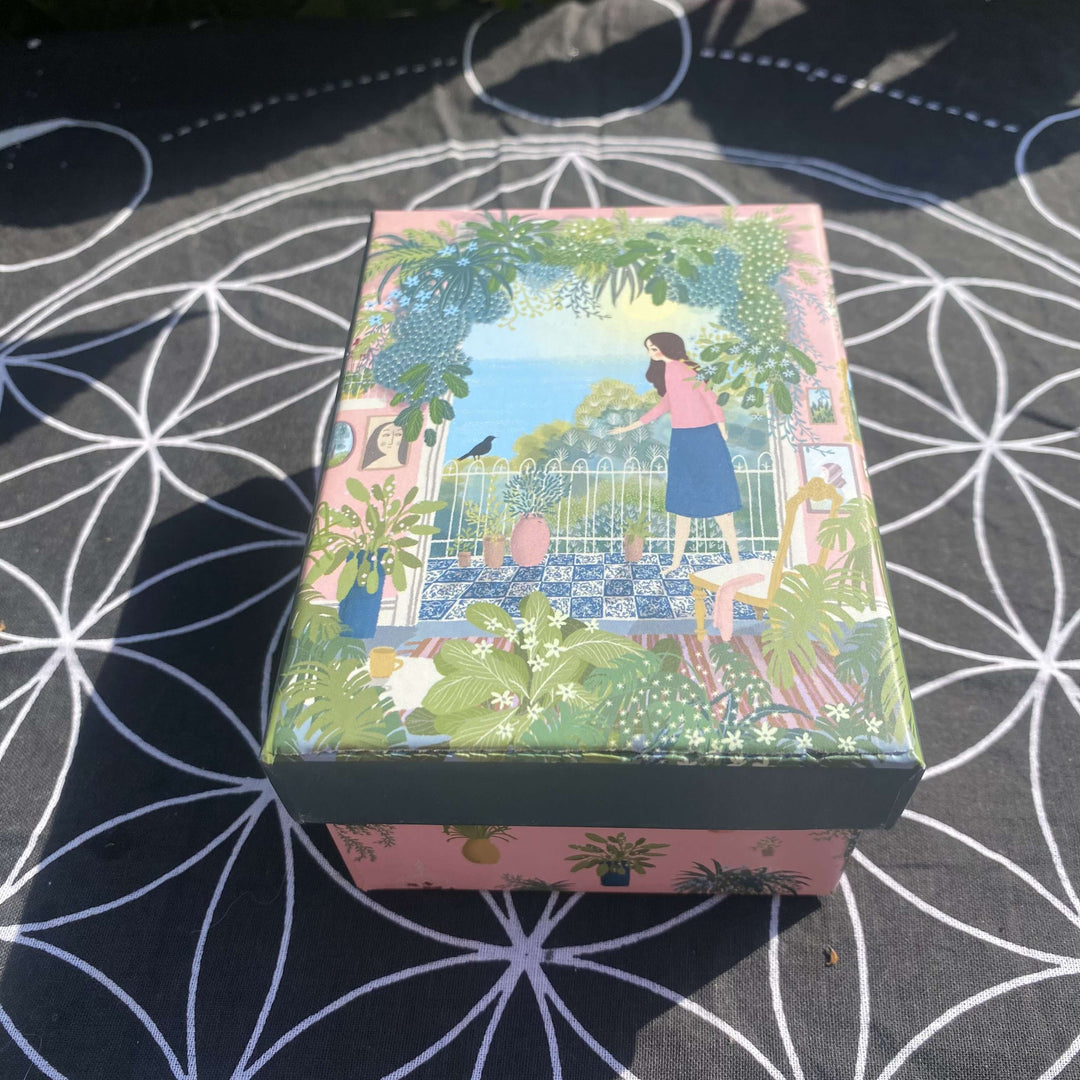 A top-down view of the closed, stylish crystal gift box resting on a black and white grid mat under bright daylight. The decorative lid features a colorful, cozy illustration of a serene garden patio scene with a figure overlooking blooming flowers.
