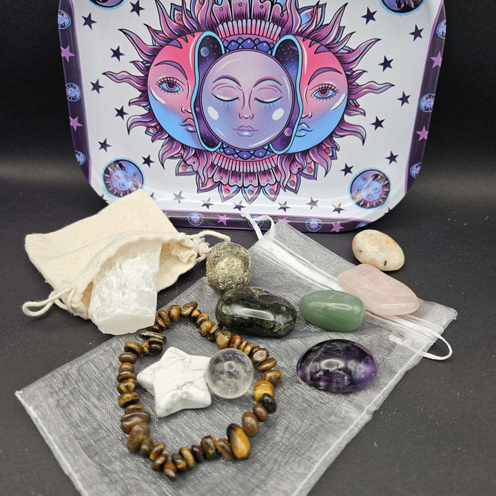 The complete Ultimate Sanctuary Kit package showing a brown tiger eye stretch bracelet and a carved howlite star. The custom-designed rectangular metal tin box stands behind the stones, featuring a vibrant psychedelic pink and purple moon face motif. 