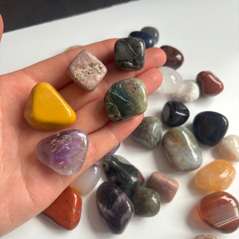 A hand tilted to present a larger assortment of pocket-sized imperfect crystal tumble stones indoors, illustrating a diverse mix of mineral tones, natural clouding, and slight surface flaws from the Quirky Corner collection.