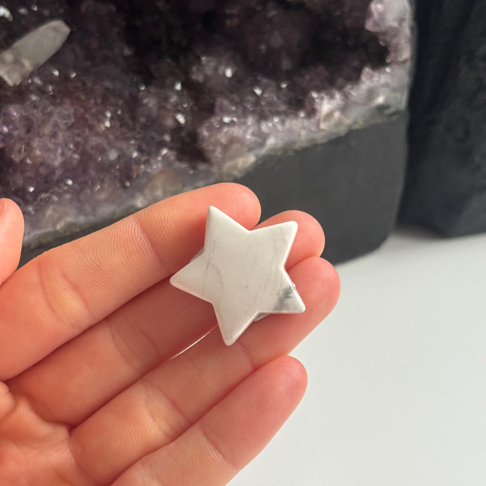 An open hand cradling the tiny white howlite pocket star carving at a mid-range distance indoors, demonstrating its compact 3cm physical sizing scale and unique marble-like porcelain finish next to a large amethyst cluster.