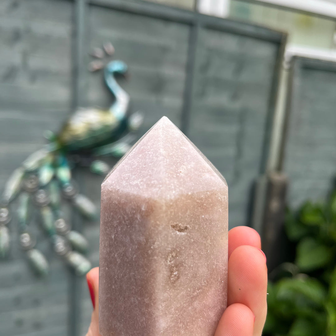 A close-up view focusing on the upper facets and pointed tip of the pink crystal generator tower held in a hand. The shot openly reveals the authentic, slight chip at its very tip, celebrating its natural geological journey from the ground straight to a cosy home sanctuary.
