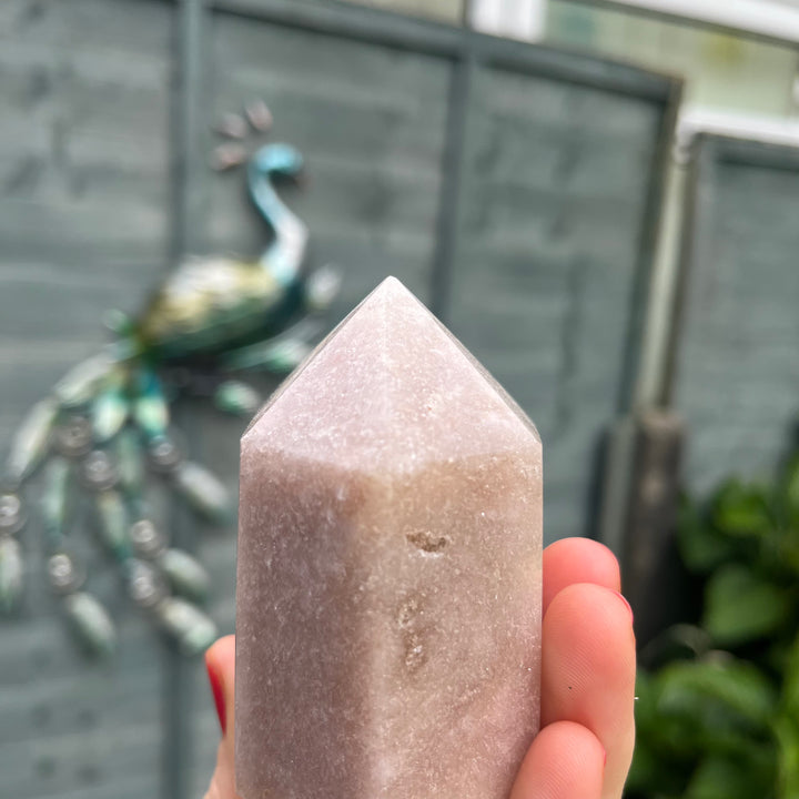 A close-up view focusing on the upper facets and pointed tip of the pink crystal generator tower held in a hand. The shot openly reveals the authentic, slight chip at its very tip, celebrating its natural geological journey from the ground straight to a cosy home sanctuary.