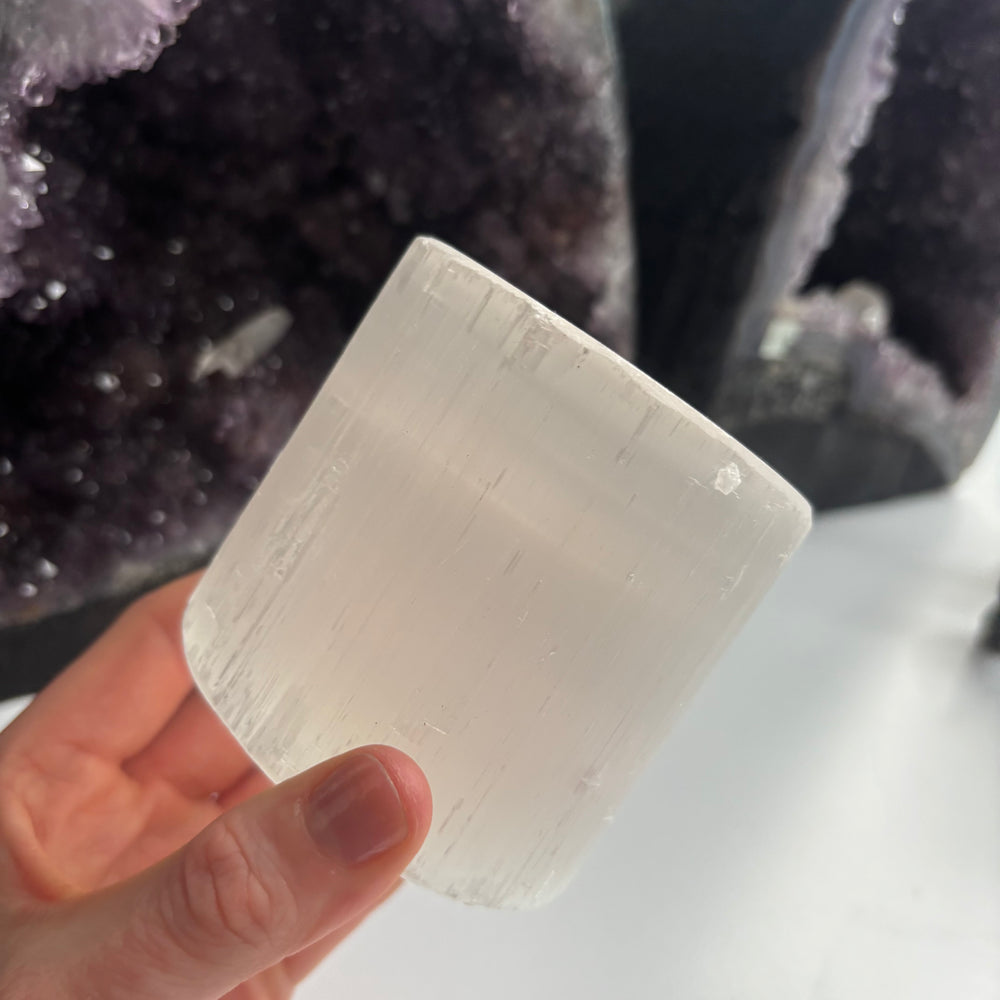 A hand holding the 8cm imperfect selenite candle holder at a distance indoors, illustrating its solid block profile, pearlescent wood-grain texture, and minor physical marks from the Quirky Corner clearance collection.