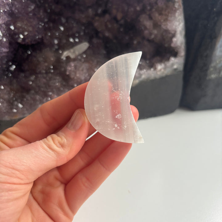 An open hand cradling the 5cm imperfect selenite crescent moon crystal at a mid-range distance indoors, demonstrating its cosy sizing scale and translucent satin white texture next to a large amethyst cluster backdrop.