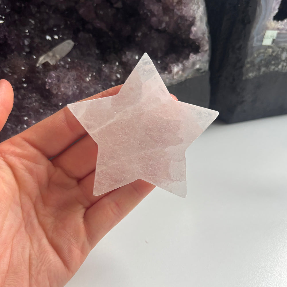 An open hand cradling the 8cm imperfect selenite crystal star carving at a mid-range distance indoors, showcasing its generous sizing scale and translucent satin white composition next to dark amethyst clusters.