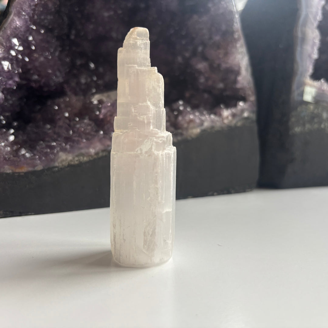 A mid-range standalone shot of the 10cm tall imperfect selenite crystal tower standing on a white table layout, illustrating its organic asymmetrical structure and ice-like raw presence from the Quirky Corner collection.