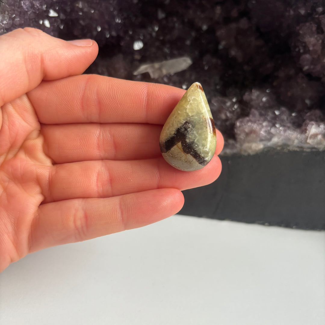 An open hand cradling the pocket-sized imperfect septarian jasper tear drop carving vertically indoors, demonstrating its lifestyle scale and unique earth-born crystalline textures next to a large cluster of raw amethyst points.
