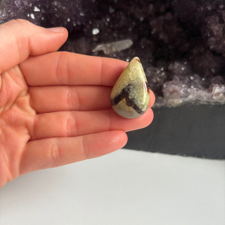 An open hand cradling the pocket-sized imperfect septarian jasper tear drop carving vertically indoors, demonstrating its lifestyle scale and unique earth-born crystalline textures next to a large cluster of raw amethyst points.