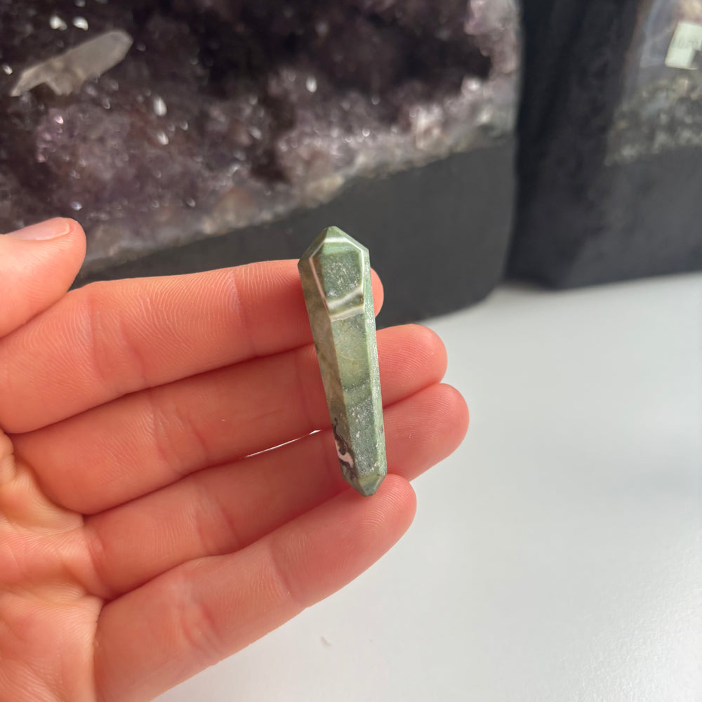 An open hand cradling the mini serpentine double-pointed crystal wand vertically indoors, demonstrating its compact 4cm physical sizing scale next to a collection of dark amethyst crystal clusters.
