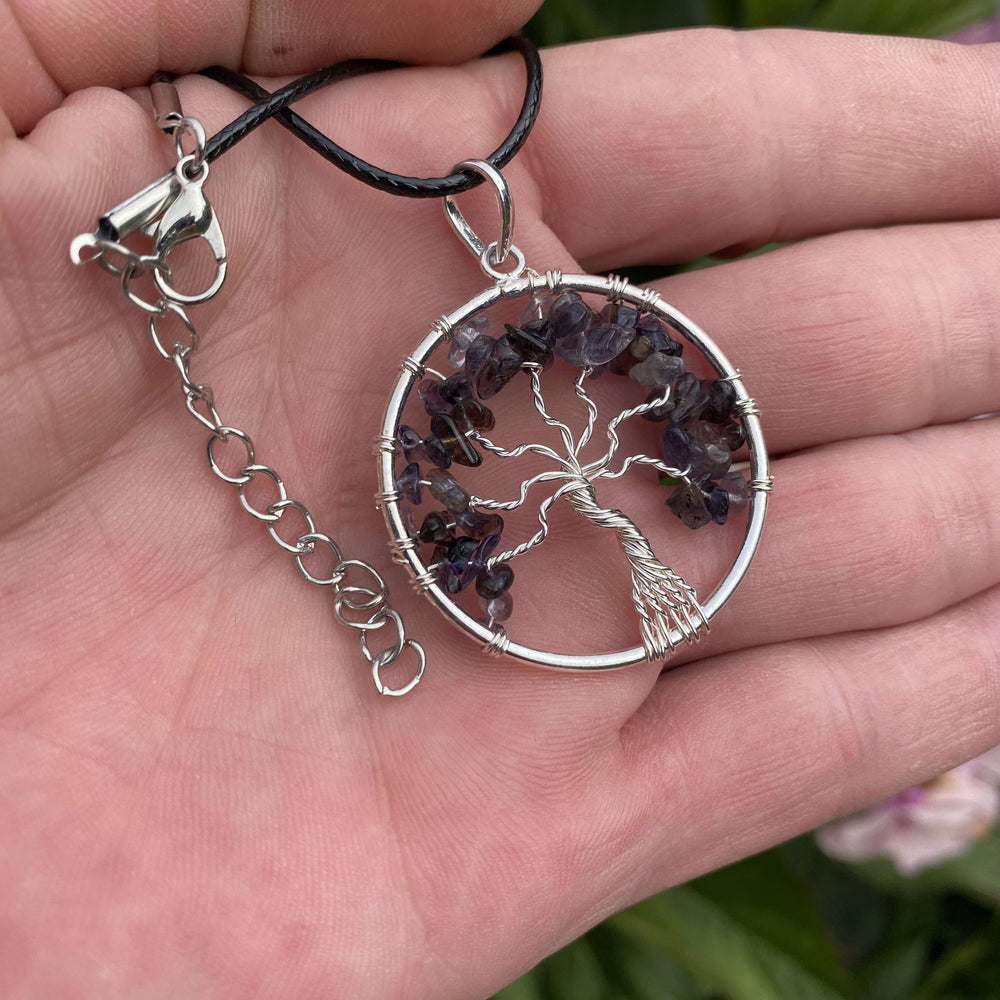 A silver tree of life charm necklace with purple gemstone accents held in an open hand, showing the black cord attached to a silver lobster clasp and extension chain.
