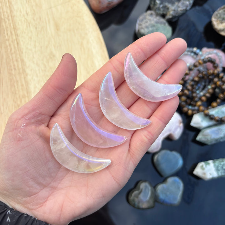 A hand presenting four iridescent aura rose quartz crescent moon shapes lined up in an open palm outdoors, illustrating the physical sizing scale, delicate veining, and metallic pastel flashes of these celestial tokens.