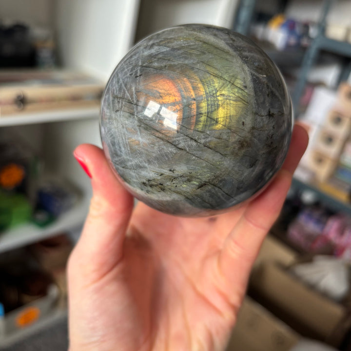 A hand presenting the hefty labradorite crystal ball, showcasing subtle shifting layers of pale green and gold schiller across its dark mineral base, perfect for styling on an intention altar or workspace layout.
