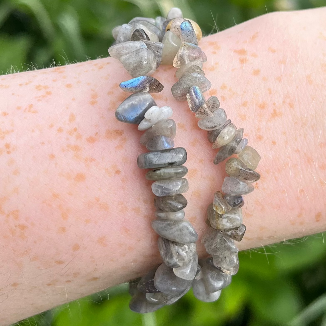 A close-up view of a freckled wrist wearing two layered labradorite crystal chip bracelets outdoors, highlighting the polished grey gemstone chips that catch the sunlight to reveal vibrant flashes of electric blue, green, and gold against a blurred garden backdrop for Third Eye and Crown Chakra alignment.
