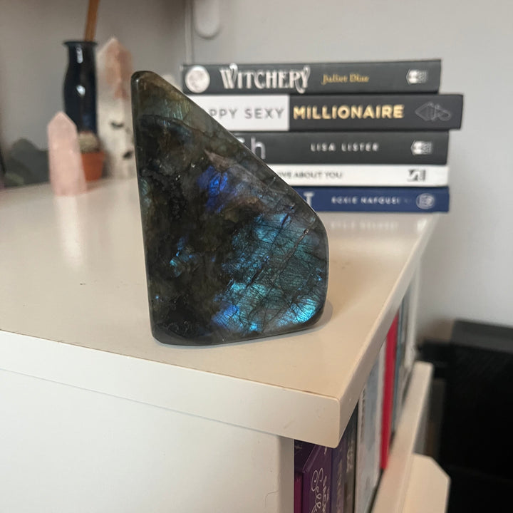 A polished labradorite crystal freeform organic sculpture standing unsupported on the edge of a white desk layout, revealing subtle flashes of deep indigo and gold along its smooth, slick facets for Third Eye chakra alignment.