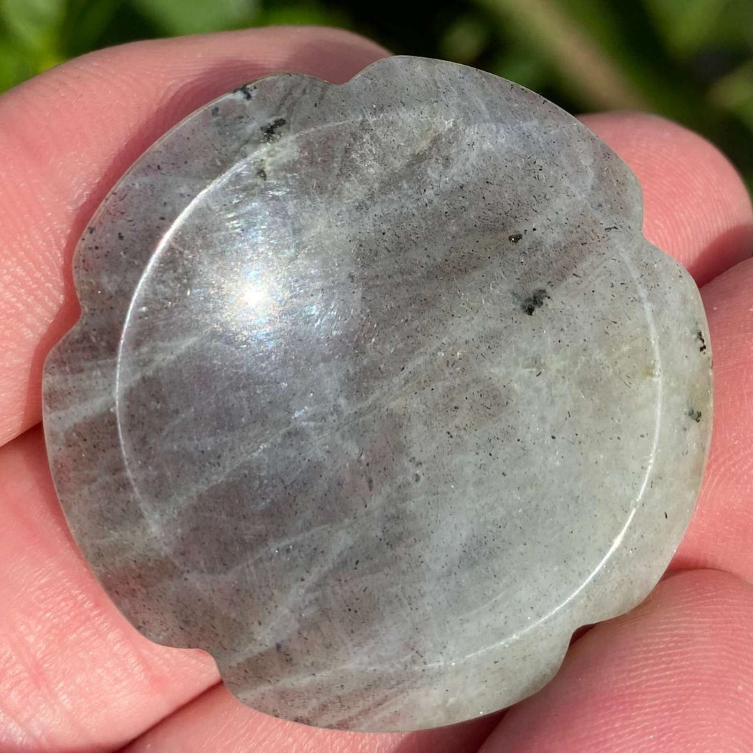 A close-up view of a carved grey labradorite flower worry stone held in an open palm. The polished stone displays subtle iridescent lines and unique earth markings across its smoothly contoured thumb indentation, reflecting themes of third eye chakra meditation.