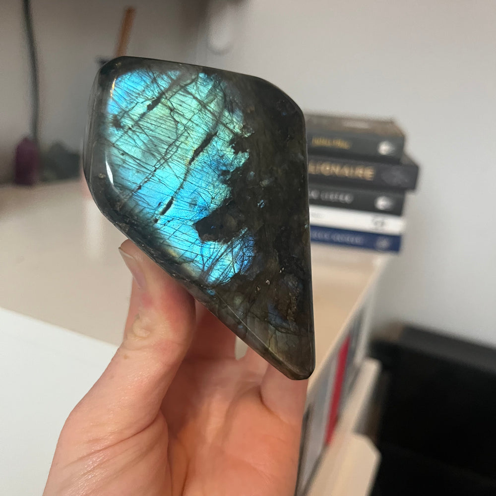 A hand tilting the 8cm tall polished labradorite freeform crystal at a sharp angle to fully capture the bright electric blue labradorescence reflecting off its glassy face, acting as a visual anchor for personal growth.
