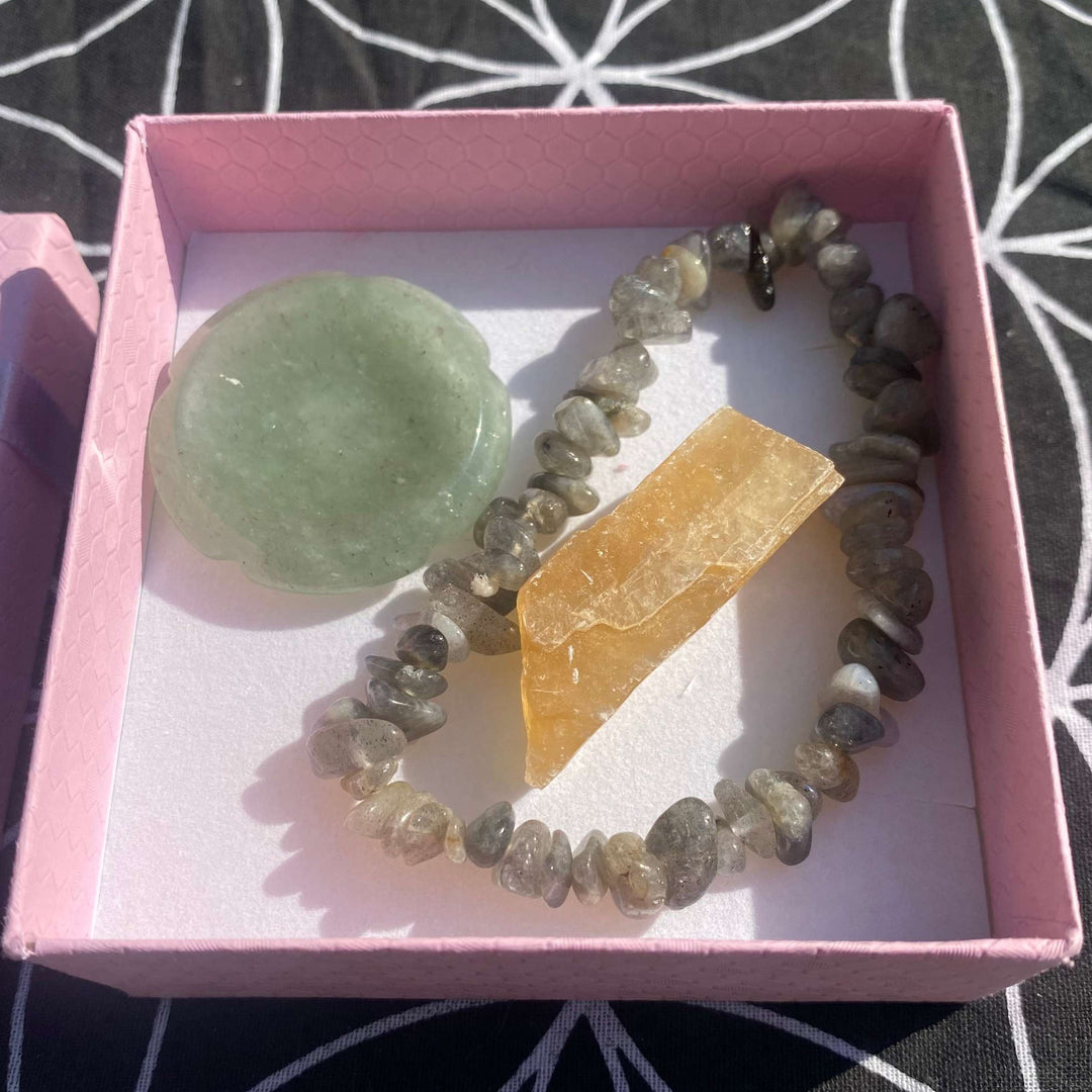 A top-down view looking into an open, square light pink box displaying the premium crystal calm gift set. Inside, an iridescent grey Labradorite chip bracelet loops around a raw, translucent golden Citrine crystal point, sitting alongside a smooth, green Aventurine flower-shaped thumbstone on a white cardboard liner.
