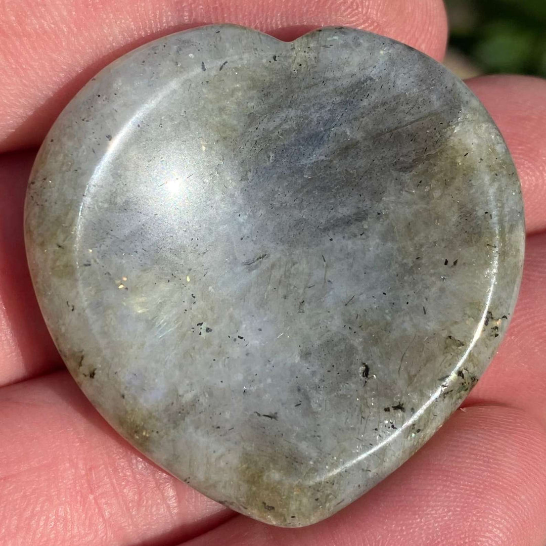 A close-up view of a polished labradorite heart worry stone resting in an open hand. The pocket stone displays its smooth, glassy grey texture and organic surface lines, highlighting a shallow thumb indentation designed for soothing third eye chakra meditation.
