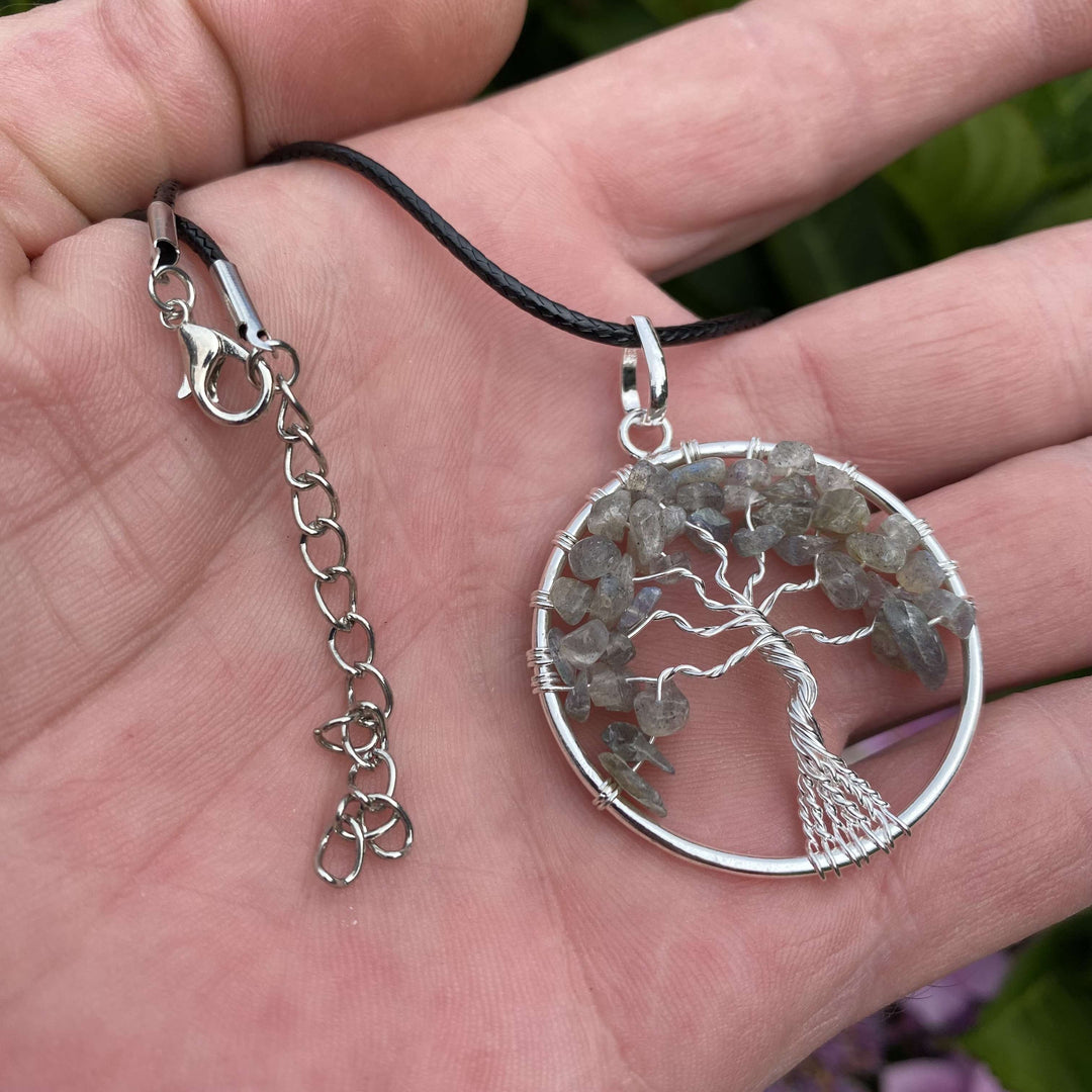 A silver circular tree of life pendant necklace featuring polished grey labradorite crystal chip leaves, resting in an open hand with a dangling silver extension chain.
