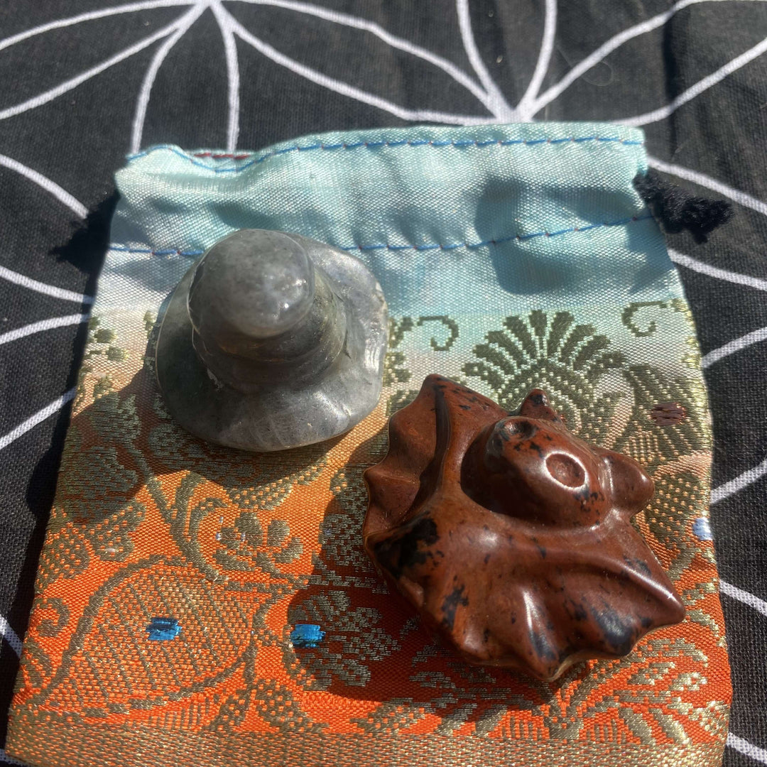 A top-down view of a grey flash labradorite witch's hat carving and a brick-red speckled snowflake obsidian bat charm on an orange and turquoise blue jacquard pouch against a sacred geometry backdrop.
