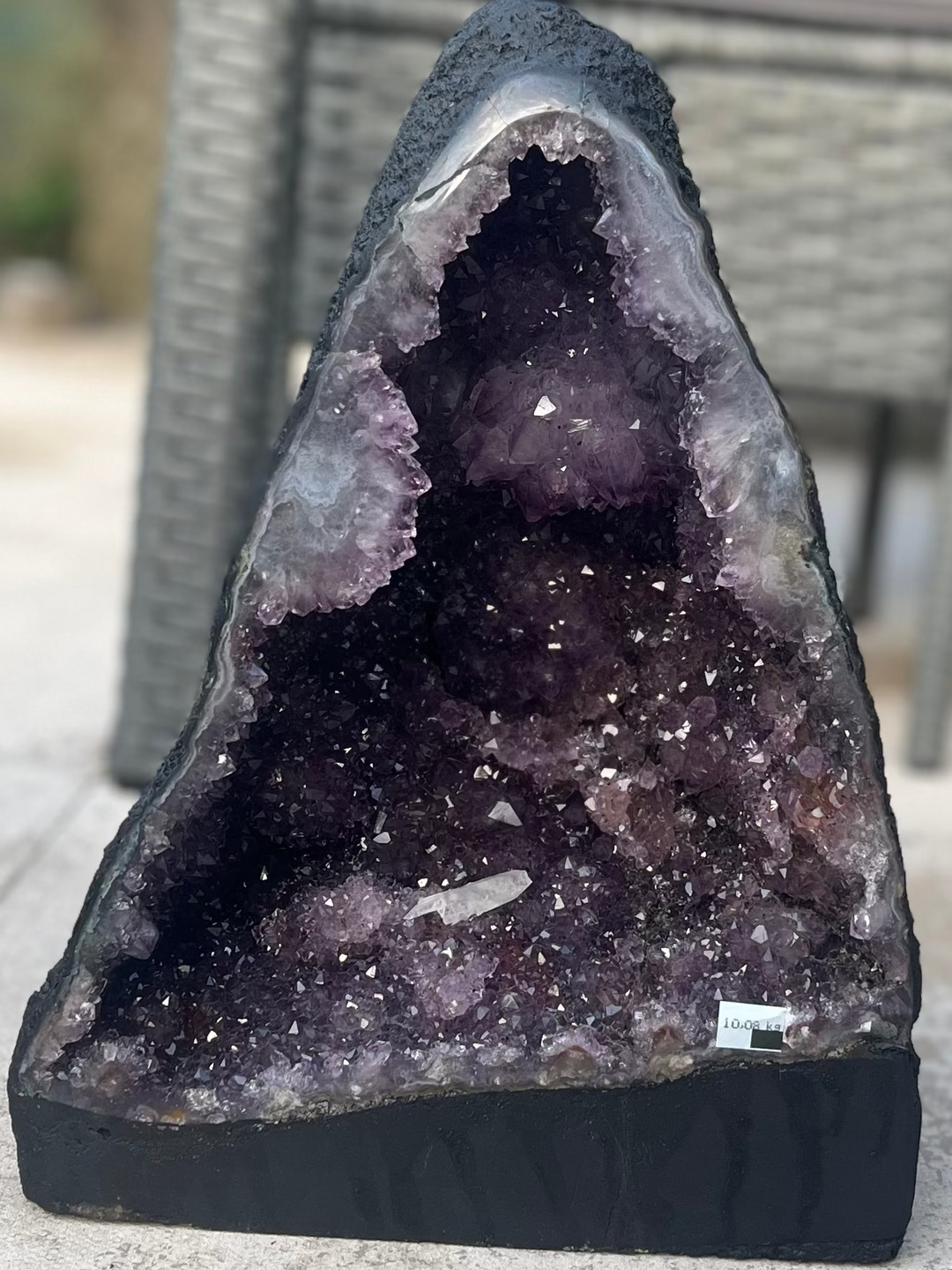 A wide shot of the wide amethyst cathedral cave resting on a stone pavement against garden chairs and table accents. The image details the distinct triangular profile and substantial base of the 11kg mineral cluster for reliable stability. 