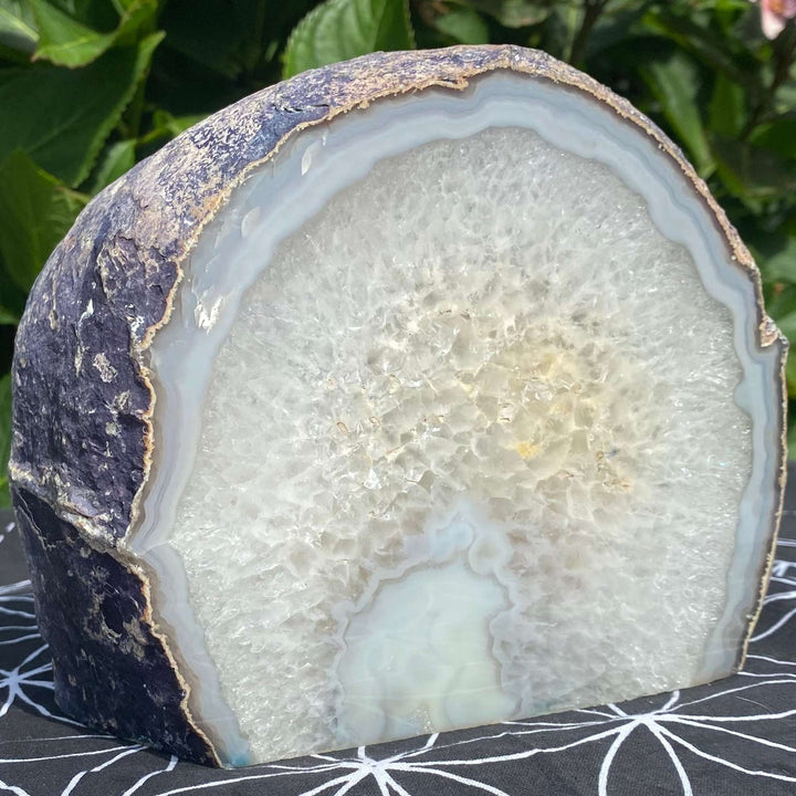 A macro close-up view of a large cut-base agate node block standing upright on a black sacred geometry mat outdoors. The polished flat face displays a wide band of icy white crystal formations ringed by a dark grey rind, revealing its grounding energy.