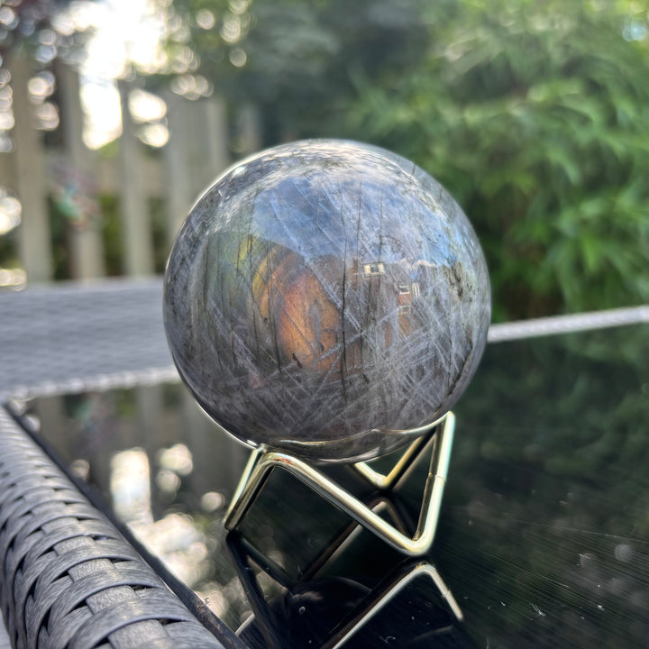 The impressive 696g large labradorite crystal sphere resting unsupported on its included three-prong gold metal stand, displayed outdoors on a black table surface to show how the symmetrical orb balances room harmony.
