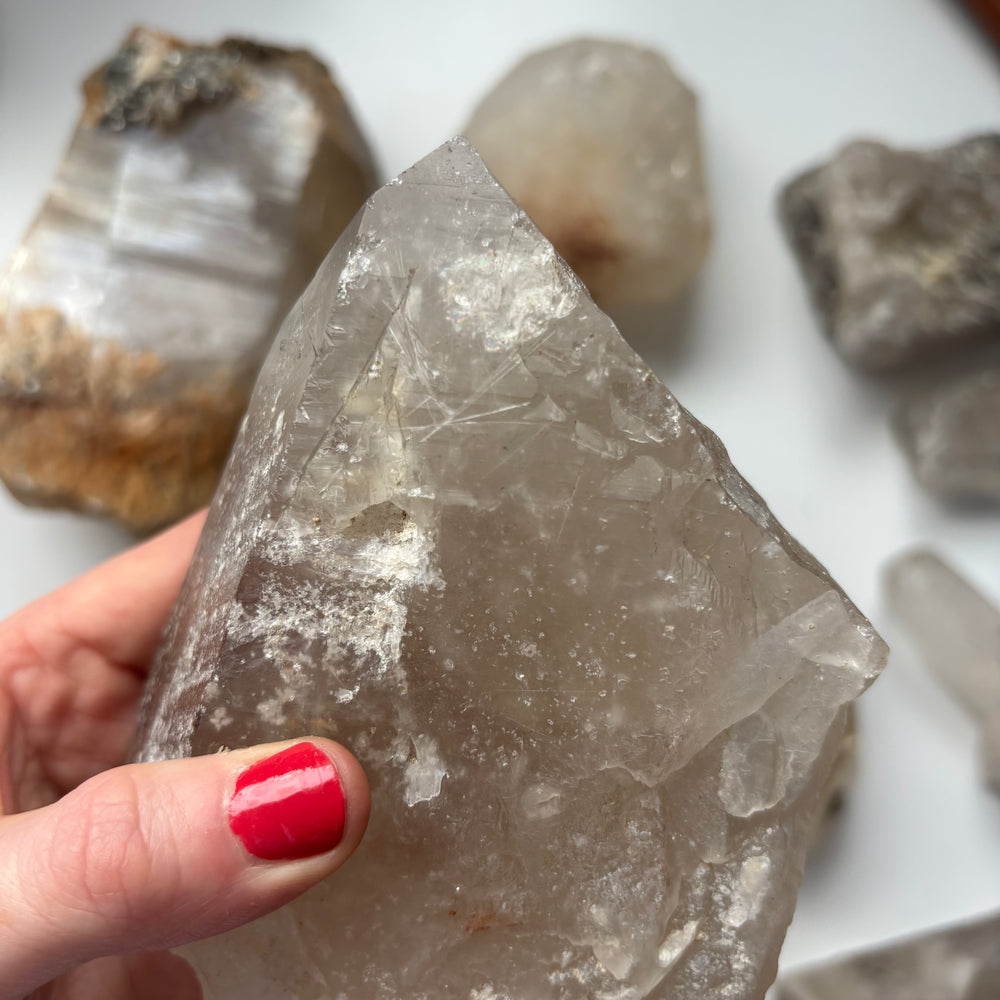 A close-up view of a hand with red nails holding the substantial rough clear quartz master healer stone, detailing the natural earth-born birthmarks and icy internal fissures that help clear away mental static and encourage life goals.